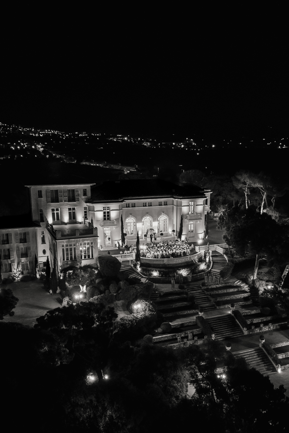 A black-and-white aerial view taken with a drone reveals Castel Bay illuminated at night, with guests gathered on the terrace for an elegant wedding reception in Provence.