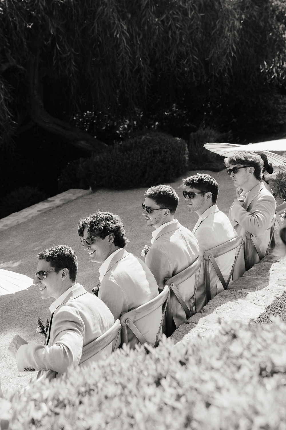 Five groomsmen in light suits and sunglasses smile while seated outdoors, waiting for the wedding ceremony at Castel Bay, South of France.