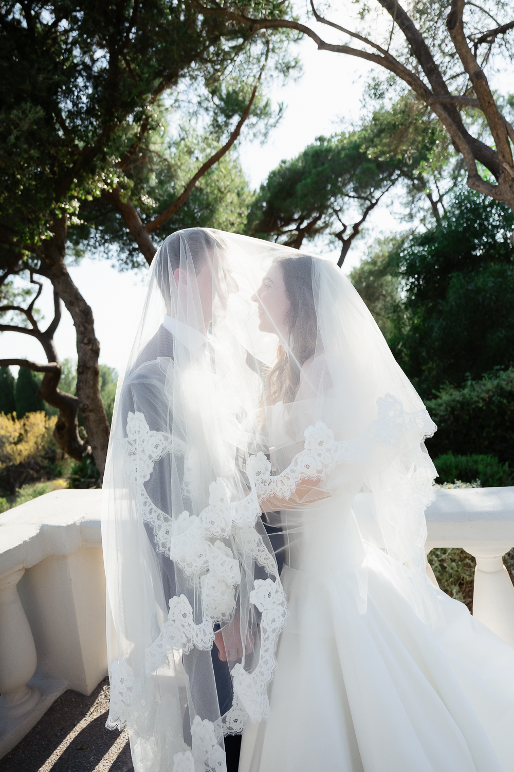 A bride and groom share a quiet moment beneath a lace-edged veil, surrounded by Mediterranean trees during a destination wedding in Castel Bay, in Provence.