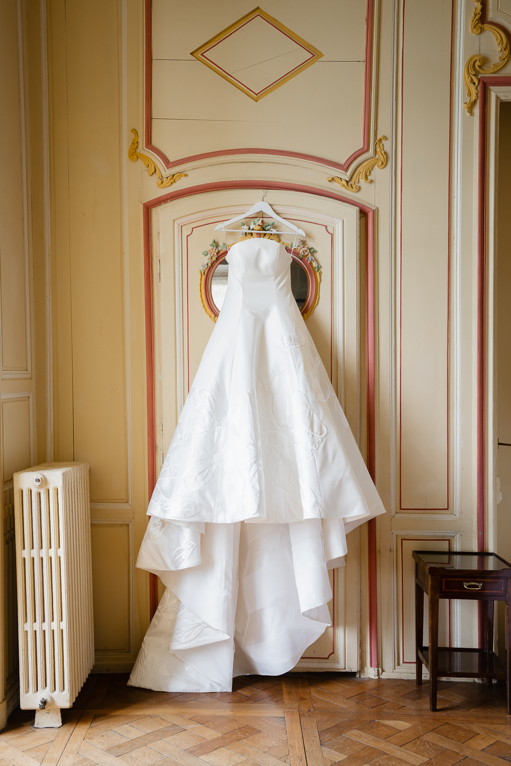 A structured wedding gown hangs in an ornate room at Château du Fey, where soft daylight reveals delicate floral embroidery and timeless bridal portrait styling.