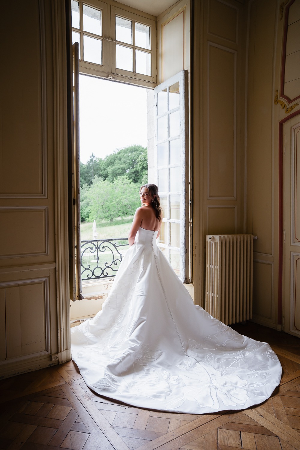 A smiling bride stands by an open window, her embroidered train flowing across the wooden floor in a quiet bridal portrait at Château du Fey.