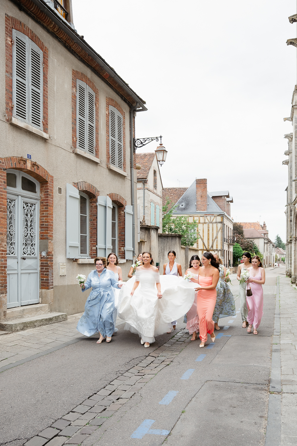 The bride walks through a quiet village street with her bridesmaids, lifting her gown as they head toward the wedding ceremony in Bourgogne.
