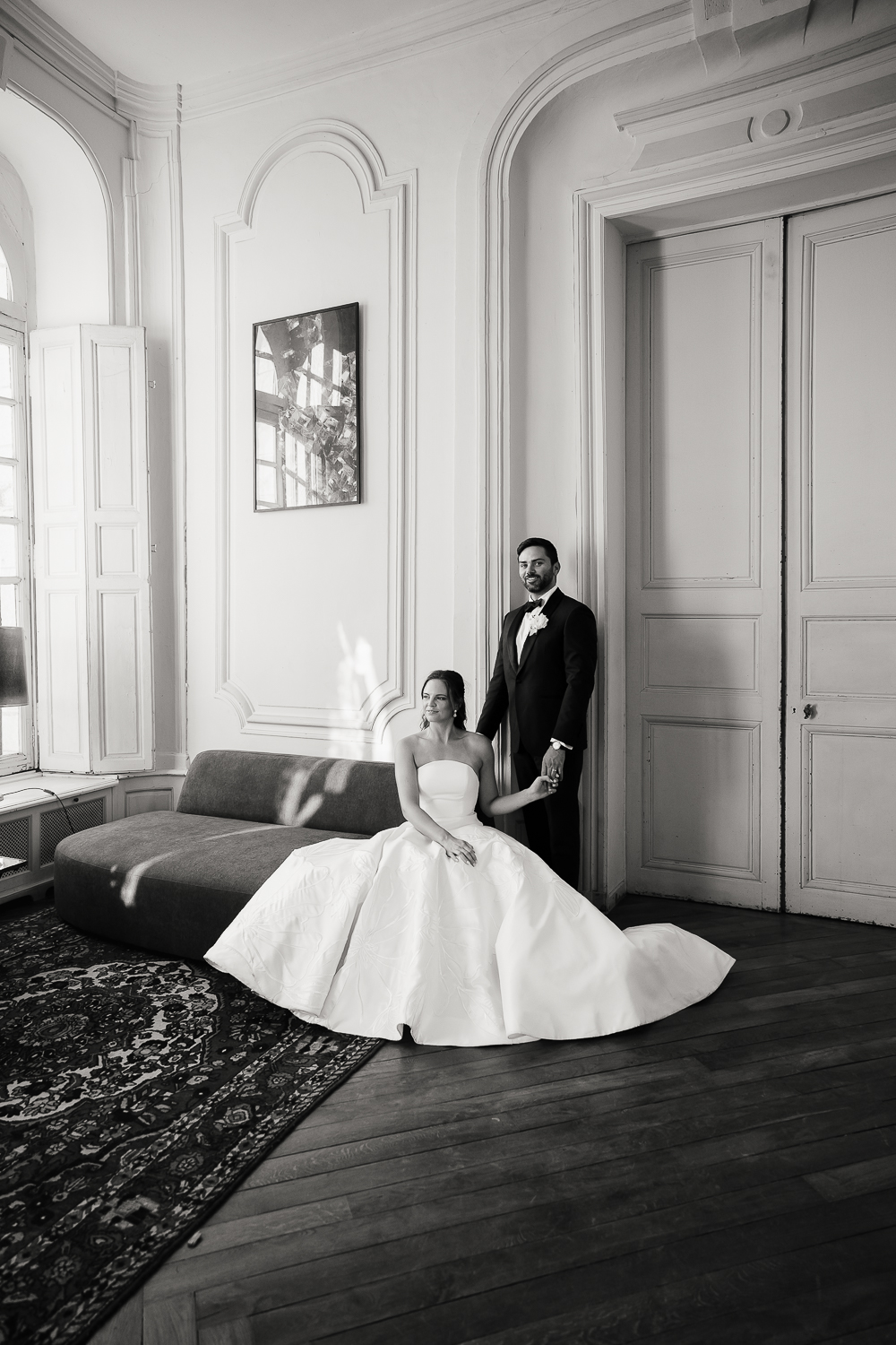 A black-and-white portrait shows the couple in an elegant château salon, framed by tall windows and classic architecture at Château du Fey.