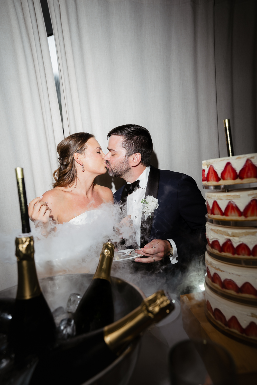 The newlyweds share a kiss beside their strawberry wedding cake, surrounded by champagne and drifting smoke during the wedding reception.