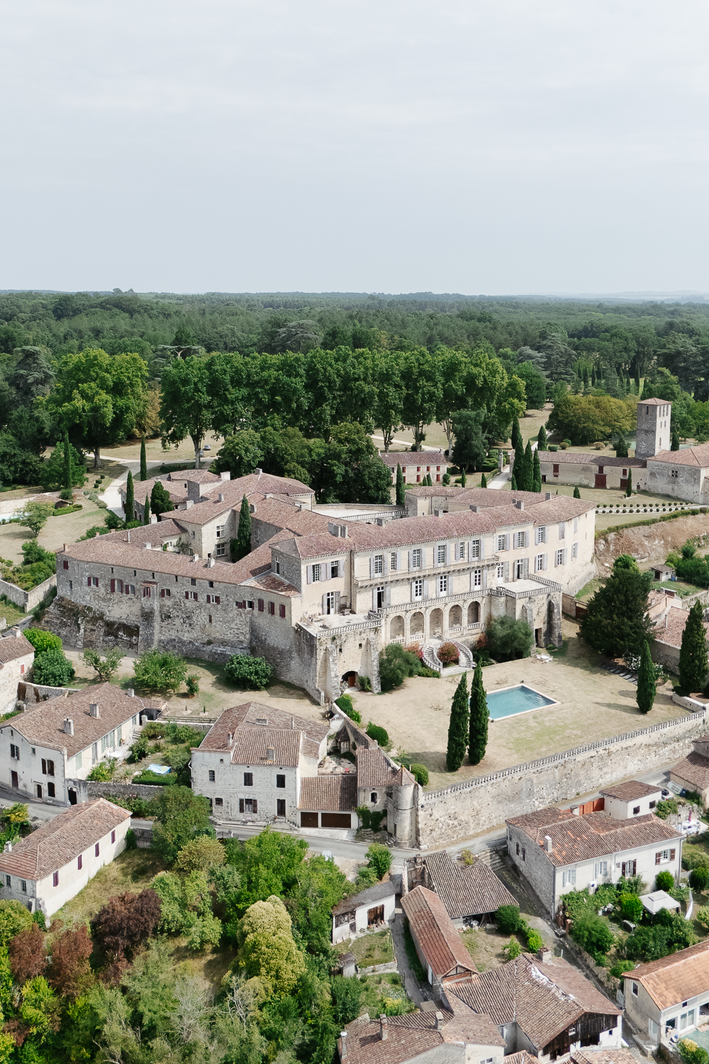 An aerial colour photograph of Château de Poudenas surrounded by cypress trees, historic stone buildings and rolling woodland, captured by Mégane Crozes, a France wedding photographer to showcase the beauty of a destination wedding venue.