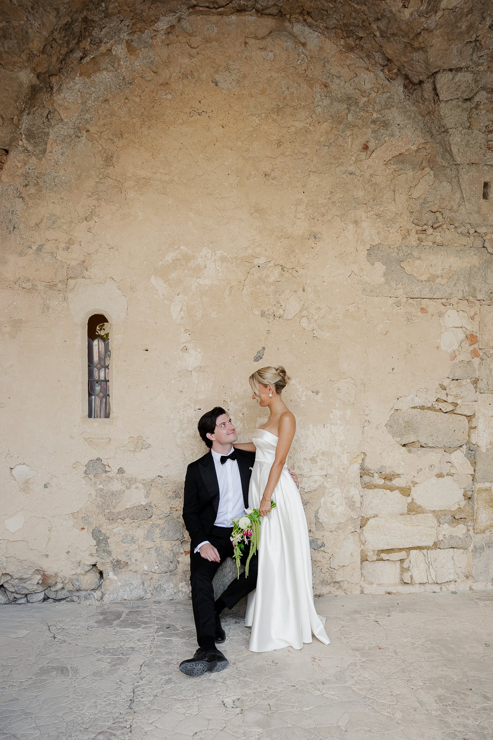 A romantic portrait of the couple beneath an ancient stone archway, where textured walls and soft natural light create a timeless editorial wedding photograph in the South of France.