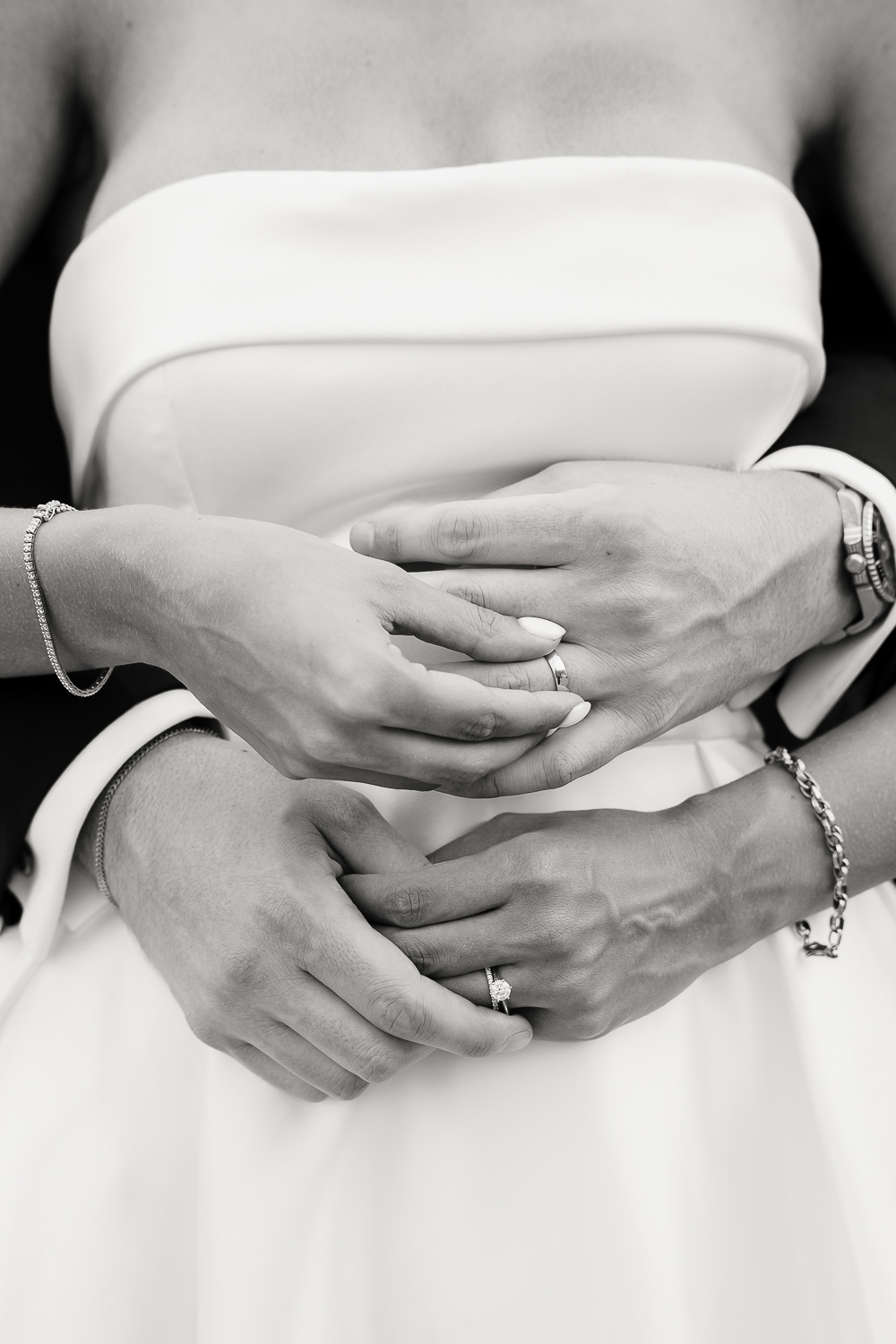 A timeless black and white close-up of the newlyweds' wedding rings resting on intertwined hands, highlighting delicate jewellery, elegant textures and the bride's minimalist satin gown.