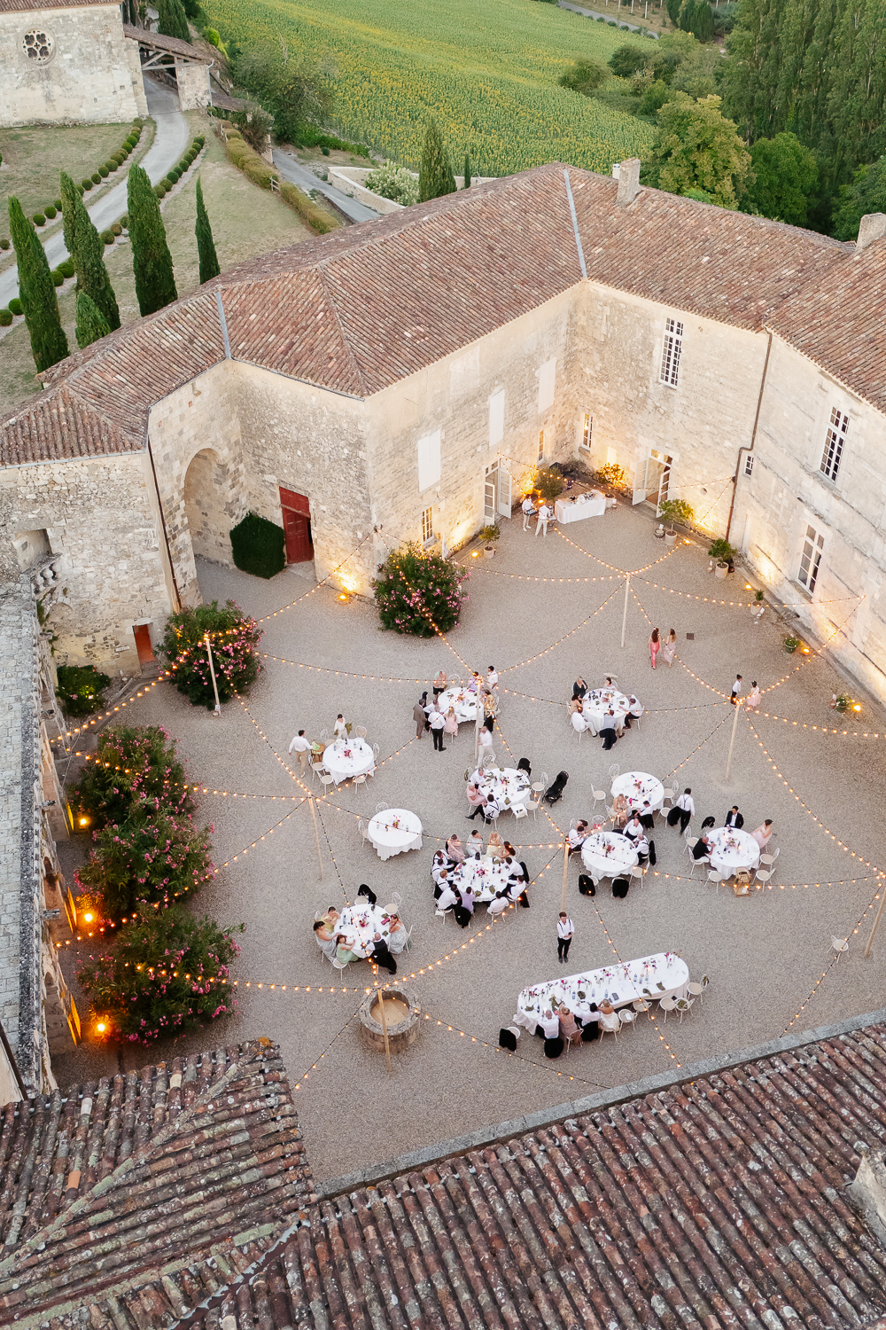 A drone view of guests dining beneath glowing string lights in the historic courtyard of Château de Poudenas, capturing the atmosphere of a refined destination wedding in France.
