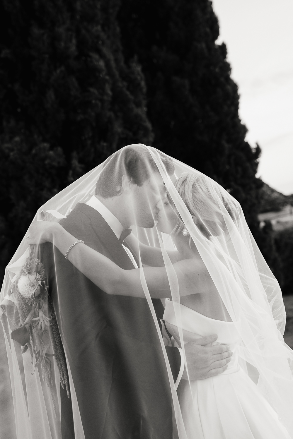 A romantic black and white portrait of the couple embracing beneath the bride's flowing veil, where soft light and translucent fabric create an intimate editorial wedding moment.