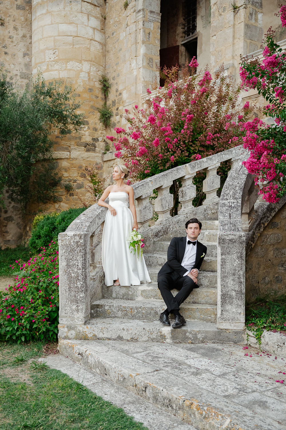 An elegant colour portrait of the bride and groom posing on the grand stone staircase of Château de Poudenas, framed by vibrant pink summer flowers and timeless French architecture.