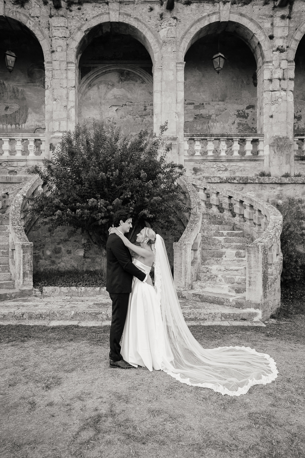 A black and white portrait captured by Mégane Crozes, featuring the newlyweds embracing in front of the château's iconic twin staircase, with the bride's long veil flowing across the lawn.