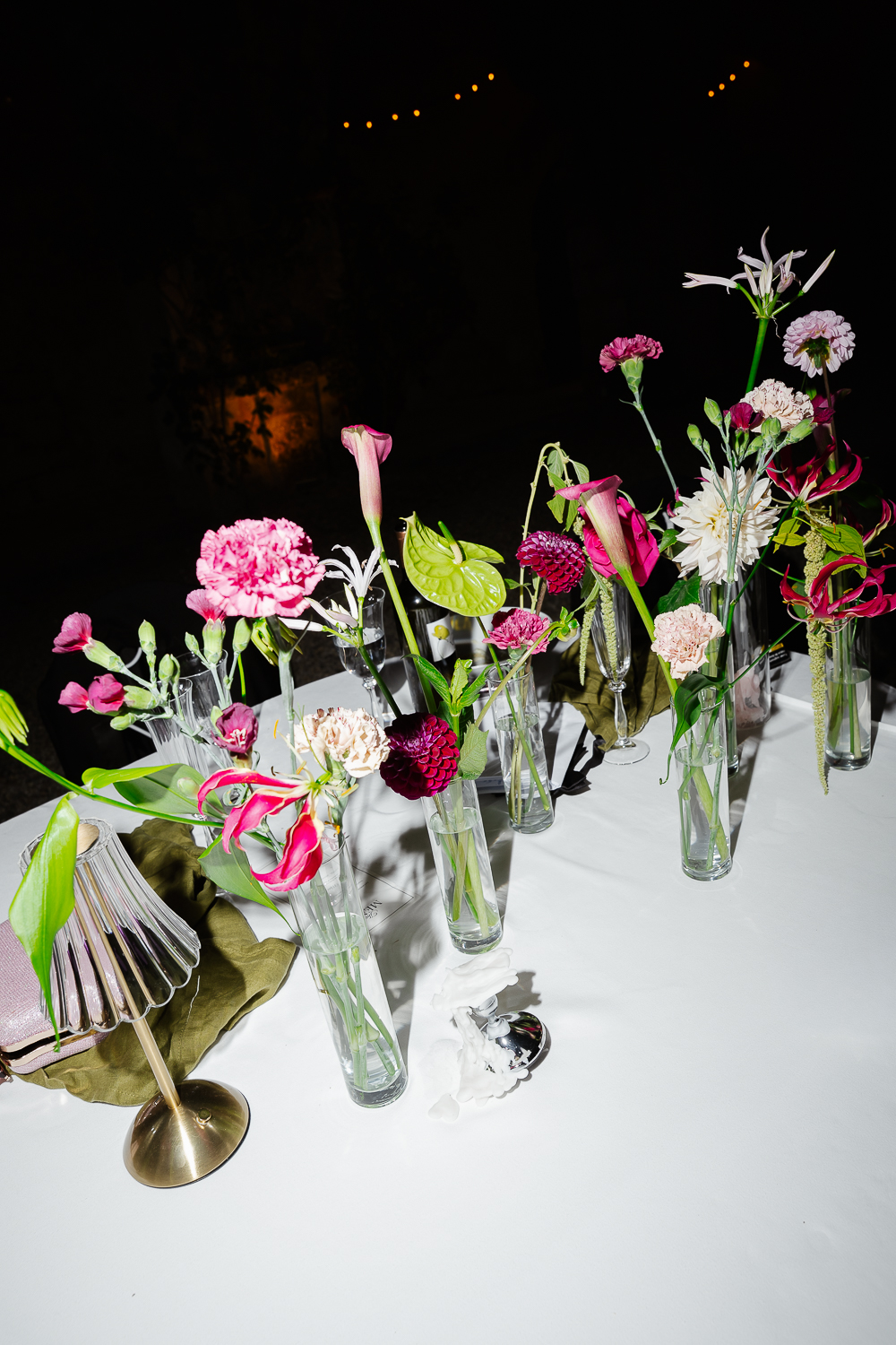 A colourful floral tablescape featuring calla lilies, hydrangeas, carnations and tropical blooms arranged in glass vases, showcasing modern wedding reception décor after sunset.