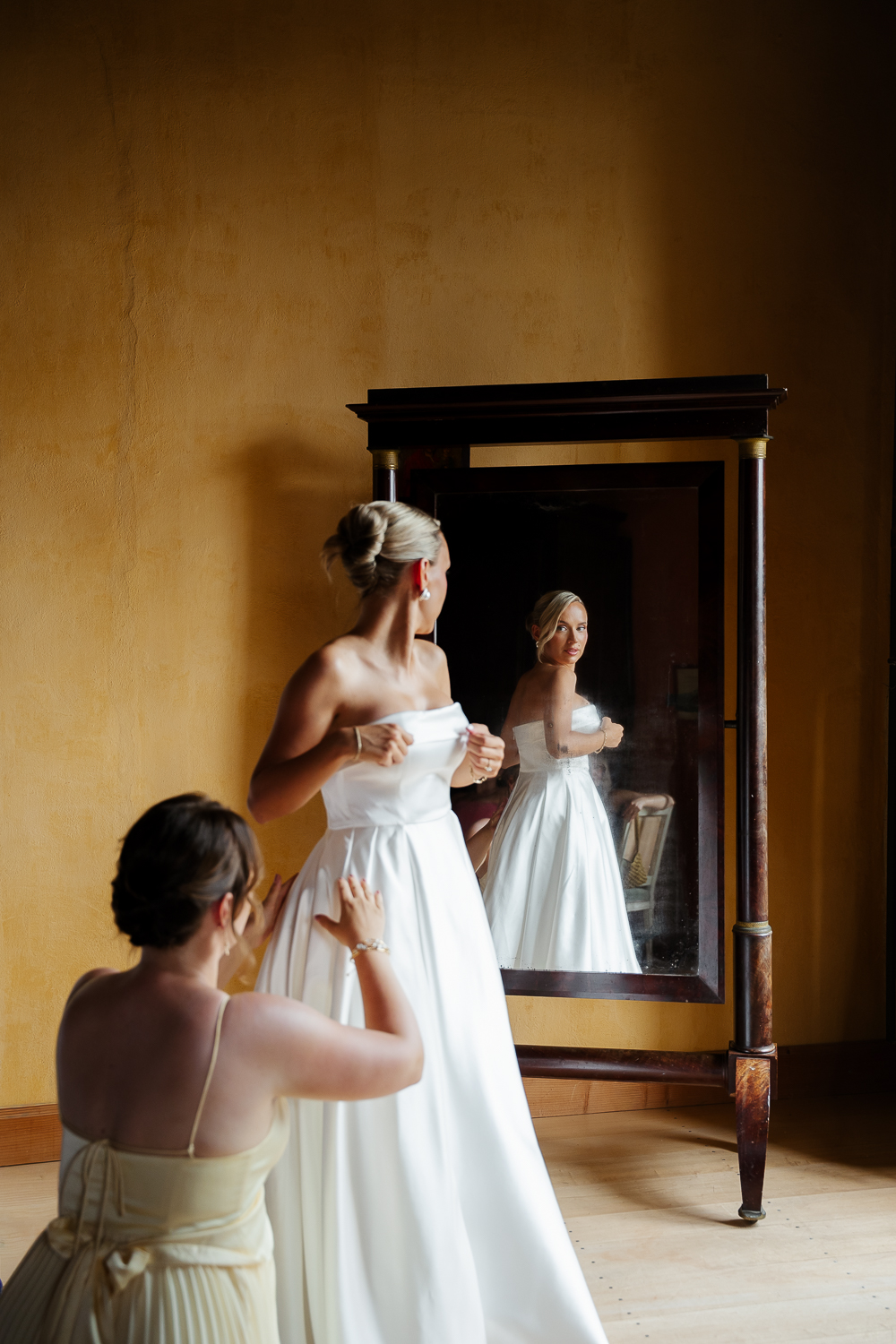 A timeless bridal preparation moment captured in colour, as the bride adjusts her elegant satin wedding gown while her bridesmaid fastens the dress. Her reflection in an antique mirror adds depth and emotion inside the historic Château de Poudenas.