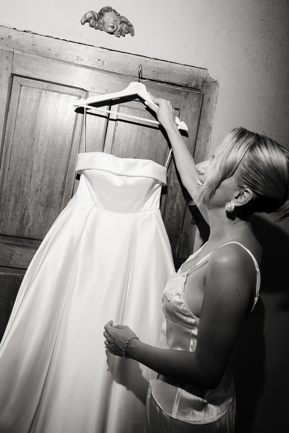 A timeless black and white portrait of a smiling bride hanging her satin wedding dress on an antique wardrobe beneath a carved angel, capturing the quiet excitement of the wedding morning.