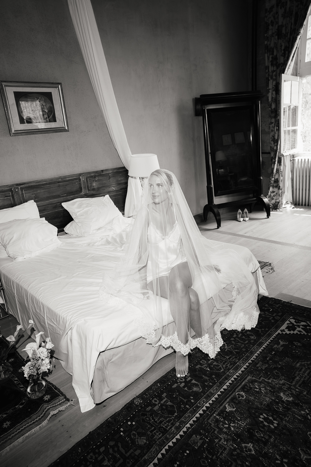 A black and white bridal portrait photographed by Mégane Crozes, where soft window light gently illuminates the veil and silk fabrics against the timeless character of the château bedroom.