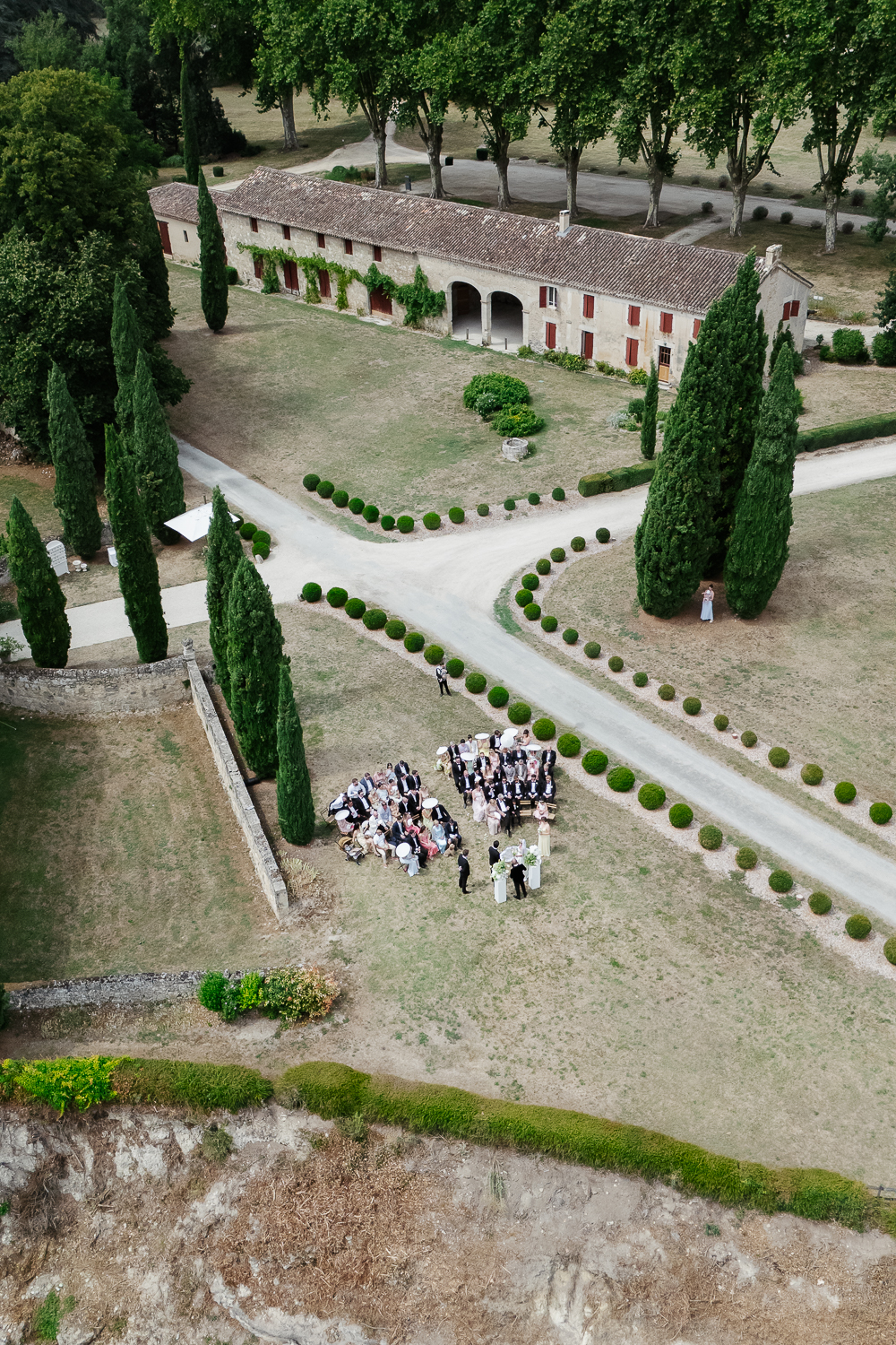 A drone photograph of a destination wedding ceremony set among cypress trees and formal gardens, with guests gathered around the couple in the French countryside at château de Poudenas.