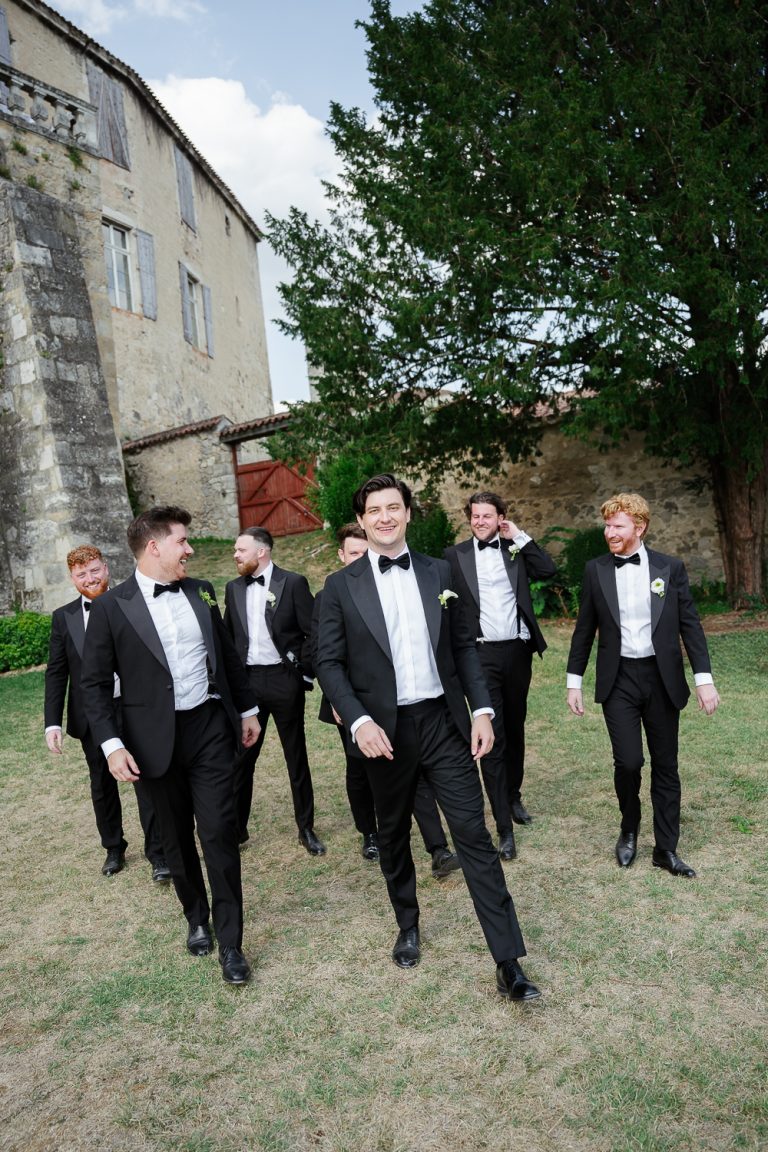The groom confidently walks across the château de Poudenas grounds with his groomsmen, their black tuxedos and genuine laughter creating a relaxed and elegant colour portrait before the ceremony.