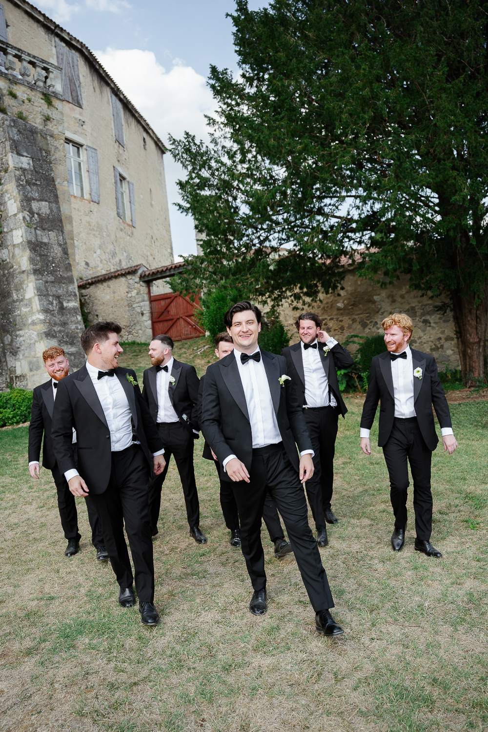 The groom confidently walks across the château de Poudenas grounds with his groomsmen, their black tuxedos and genuine laughter creating a relaxed and elegant colour portrait before the ceremony.